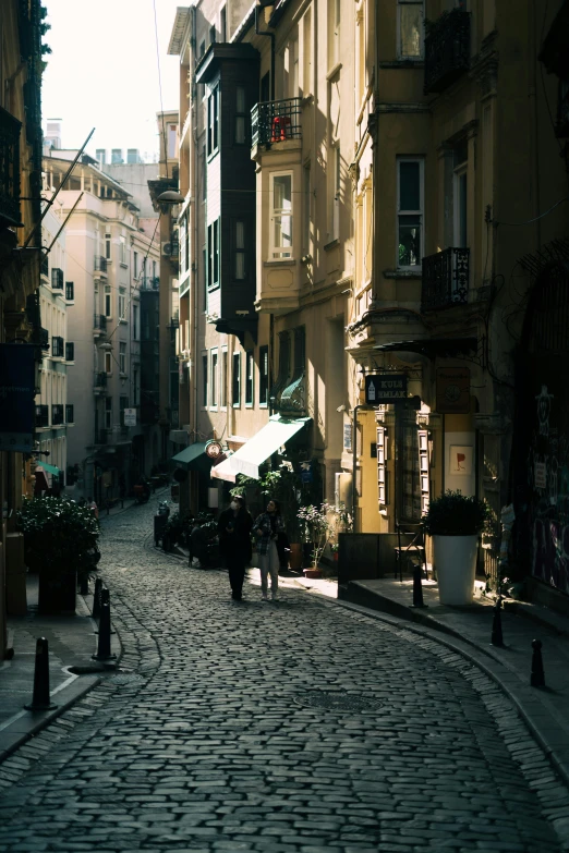 a couple of people walking down a cobblestone street, by Niyazi Selimoglu, unsplash contest winner, art nouveau, late afternoon light, fallout style istanbul, lots of shops, view from the street