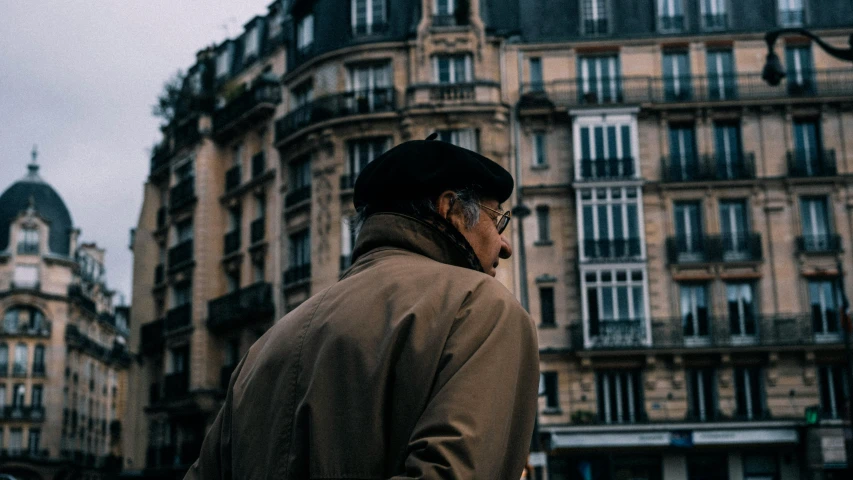 a man standing in front of a tall building, by Raphaël Collin, pexels contest winner, elderly, light brown trenchcoat, french village exterior, wearing a french beret