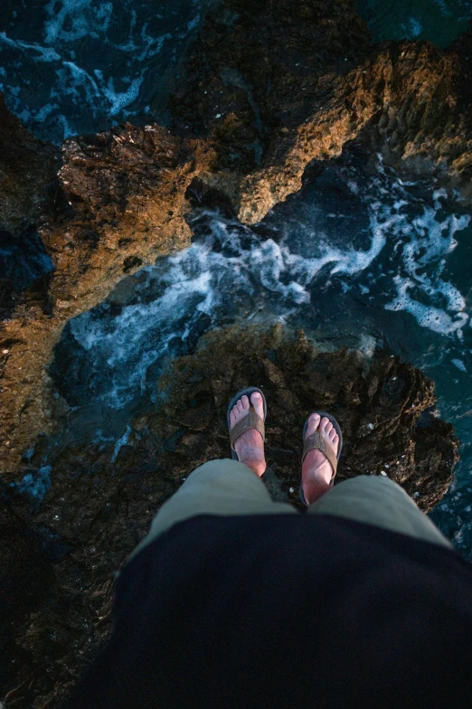 a person standing on top of a rock next to the ocean, looking down from above, flip flops, ocean floor, explore