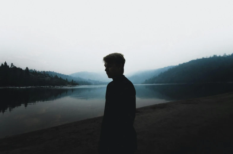 a man standing in front of a body of water, dark aesthetic, hills in the background, chillhop, profile posing