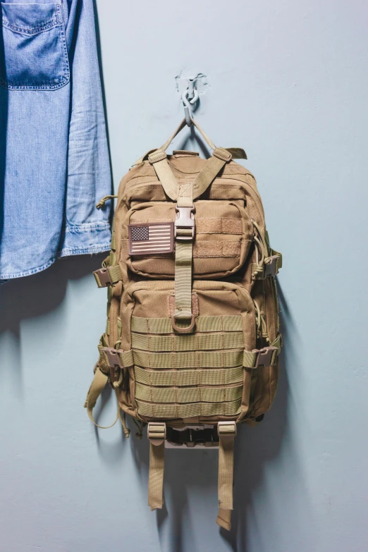 a backpack hanging on a wall next to a blue shirt, white american soldier, light tan, full product shot, square backpack