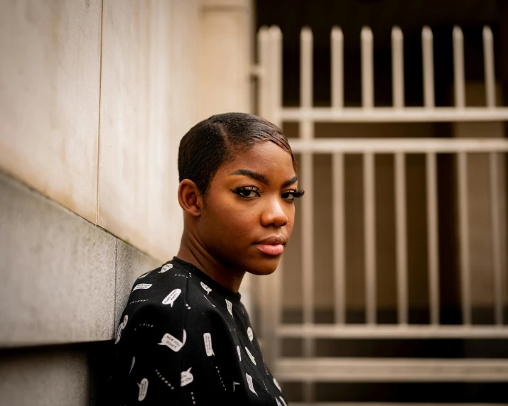 a close up of a person leaning against a wall, by Lily Delissa Joseph, short slicked - back hair, black teenage girl, proud serious expression, shot on sony a 7