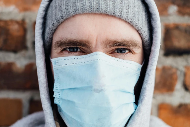 a man wearing a face mask in front of a brick wall, trending on pexels, frozen cold stare, surgical gear, thumbnail, hooded