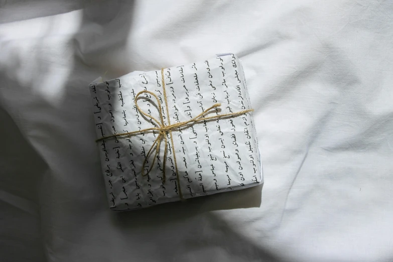 a wrapped gift sitting on top of a bed, by Alice Mason, unsplash, letterism, white poet shirt, arabic calligraphy, sustainable materials, ignant