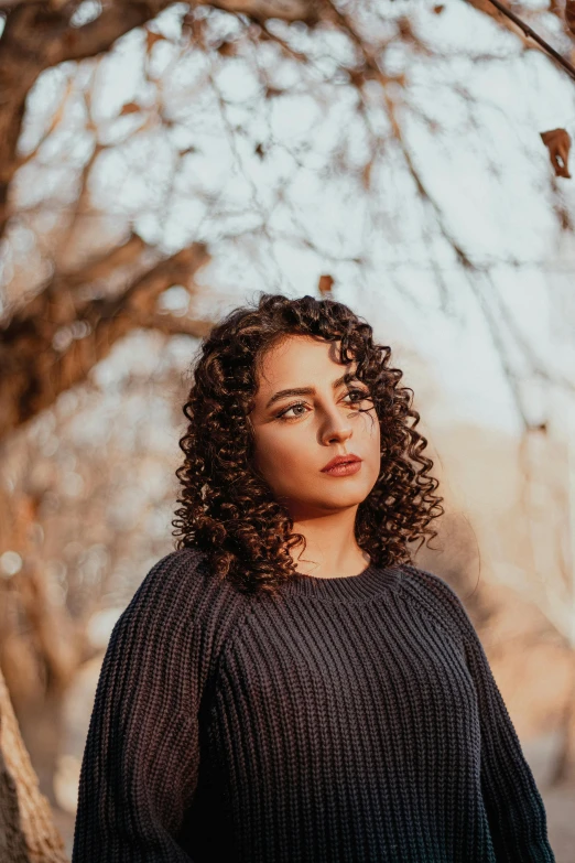 a woman with curly hair standing in front of a tree, an album cover, by irakli nadar, pexels contest winner, wearing a black sweater, looking serious, fair olive skin, sunset light