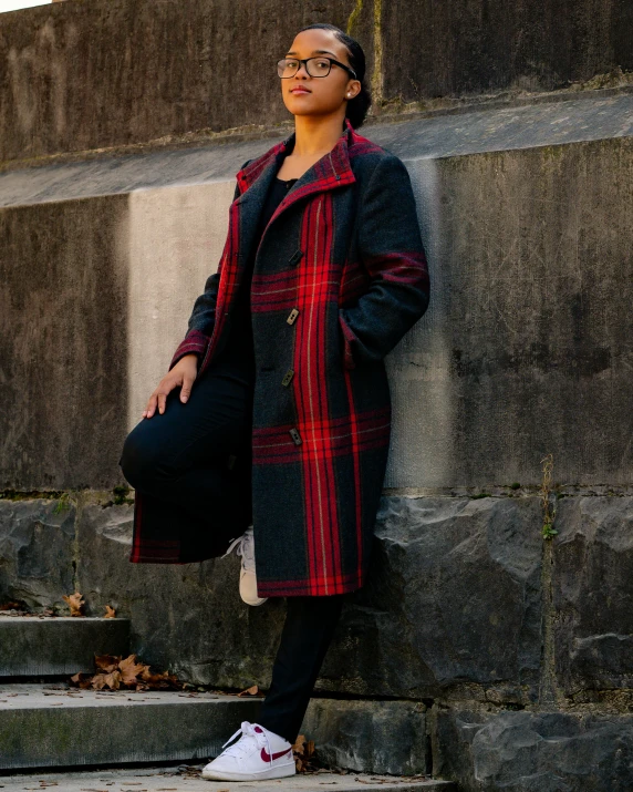 a woman leaning against a wall with a hat on her head, an album cover, by Julia Pishtar, pexels contest winner, hurufiyya, full body black and red longcoat, tartan garment, on a sidewalk of vancouver, non binary model