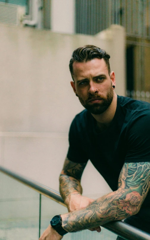 a man with tattoos sitting on a railing, promotional image, with a beard and a black shirt, anthony howe, catalog photo