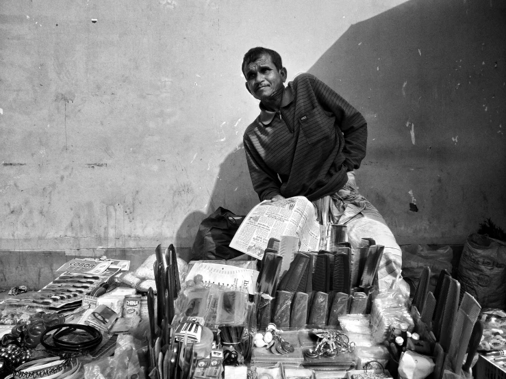 a black and white photo of a man sitting on a table, by Kailash Chandra Meher, pexels, assemblage, selling his wares, pen, with a bunch of stuff, on a sunny day