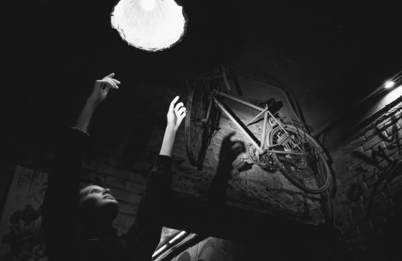 a black and white photo of a person reaching up to a light, by Brassaï, bicycle, in an attic, svetlana belyaeva, 1950s photograph