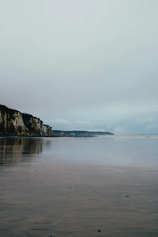 a man riding a surfboard on top of a sandy beach, a picture, inspired by Pierre Pellegrini, unsplash, romanticism, chalk cliffs above, wet relections, dunkirk, wide long view