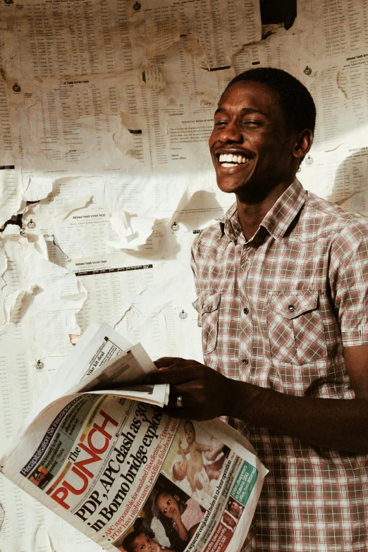 a man standing in front of a wall holding a newspaper, happening, brown skin man with a giant grin, studying in a brightly lit room, press shot, african man