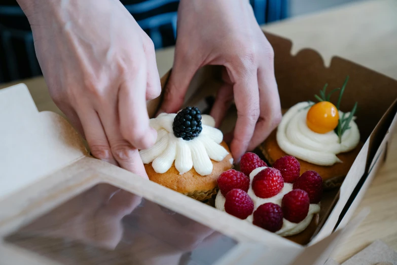 a person putting icing on a cupcake in a box, a still life, by Niko Henrichon, trending on unsplash, steamed buns, “berries, in the sun, carefully designed