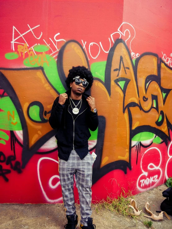 a man standing in front of a graffiti covered wall, an album cover, trending on pexels, graffiti, 2 1 savage, rapper bling jewelry, with afro, professional iphone photo