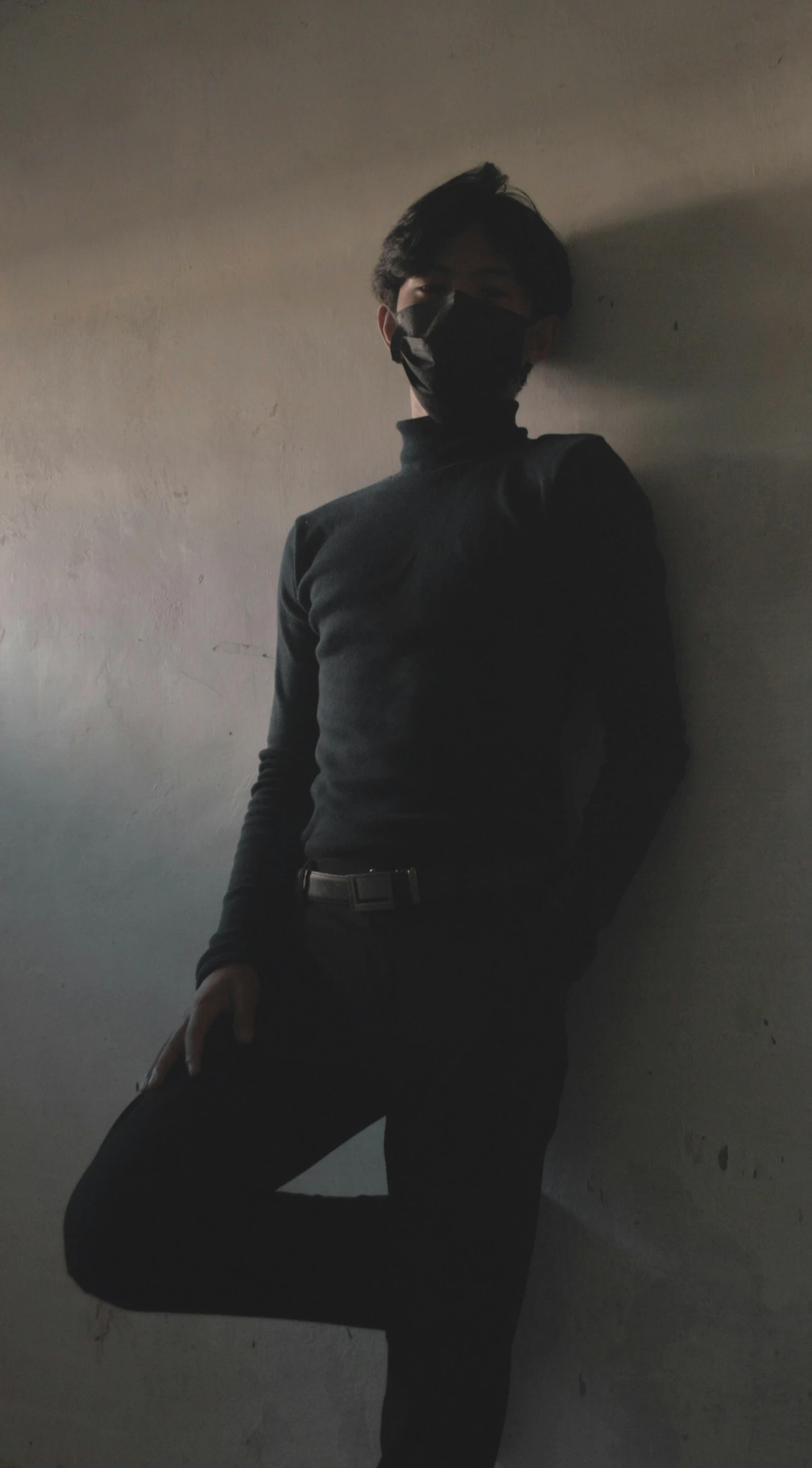 a man in a gas mask leaning against a wall, an album cover, unsplash, wearing turtleneck, charcoal color skin, low quality photo, pose 4 of 1 6