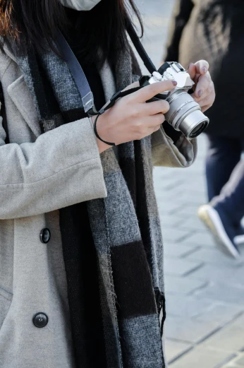 a woman standing on a sidewalk holding a camera, trending on pexels, scarf, grey and silver, a photo of a man, coat pleats