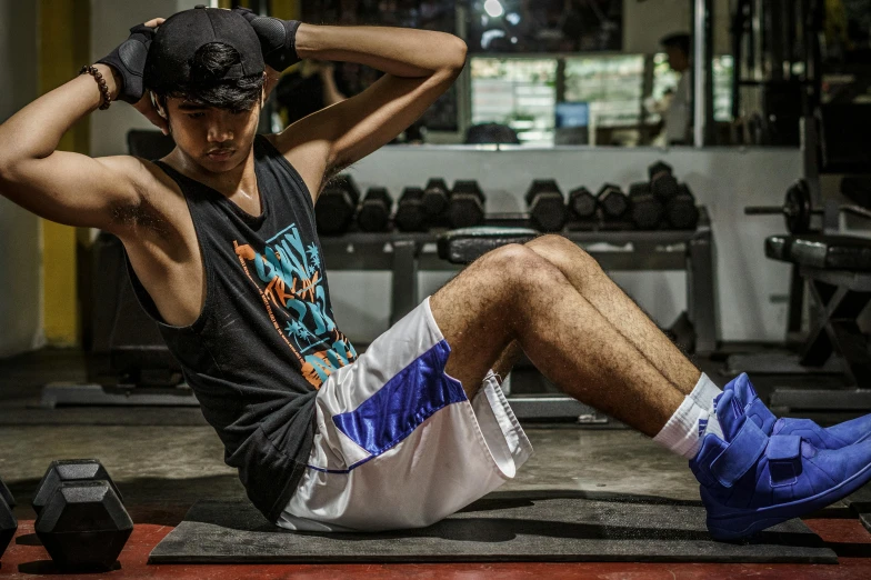 a man sitting on a mat in a gym, by Robbie Trevino, pexels contest winner, baggy clothing and hat, avatar image, thailand, physical : tinyest midriff ever