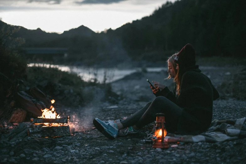 a woman sitting next to a campfire looking at her phone, a picture, by Niko Henrichon, pexels contest winner, best friends, fishing, al fresco, vibing to music