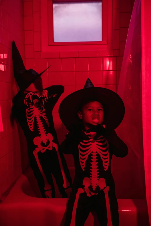 two children dressed up in halloween costumes in a bathtub, by Gwen Barnard, pexels contest winner, symbolism, red neon lights inside it, black witch hat, red room, walking over a skeleton