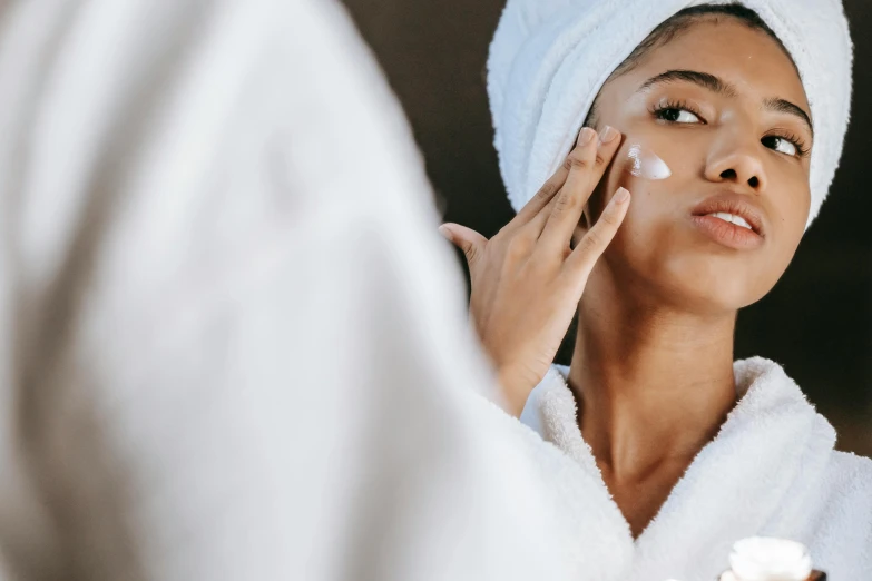 a woman putting cream on her face in front of a mirror, trending on pexels, renaissance, wearing long white robe, deep tan skin, woman holding another woman, manuka