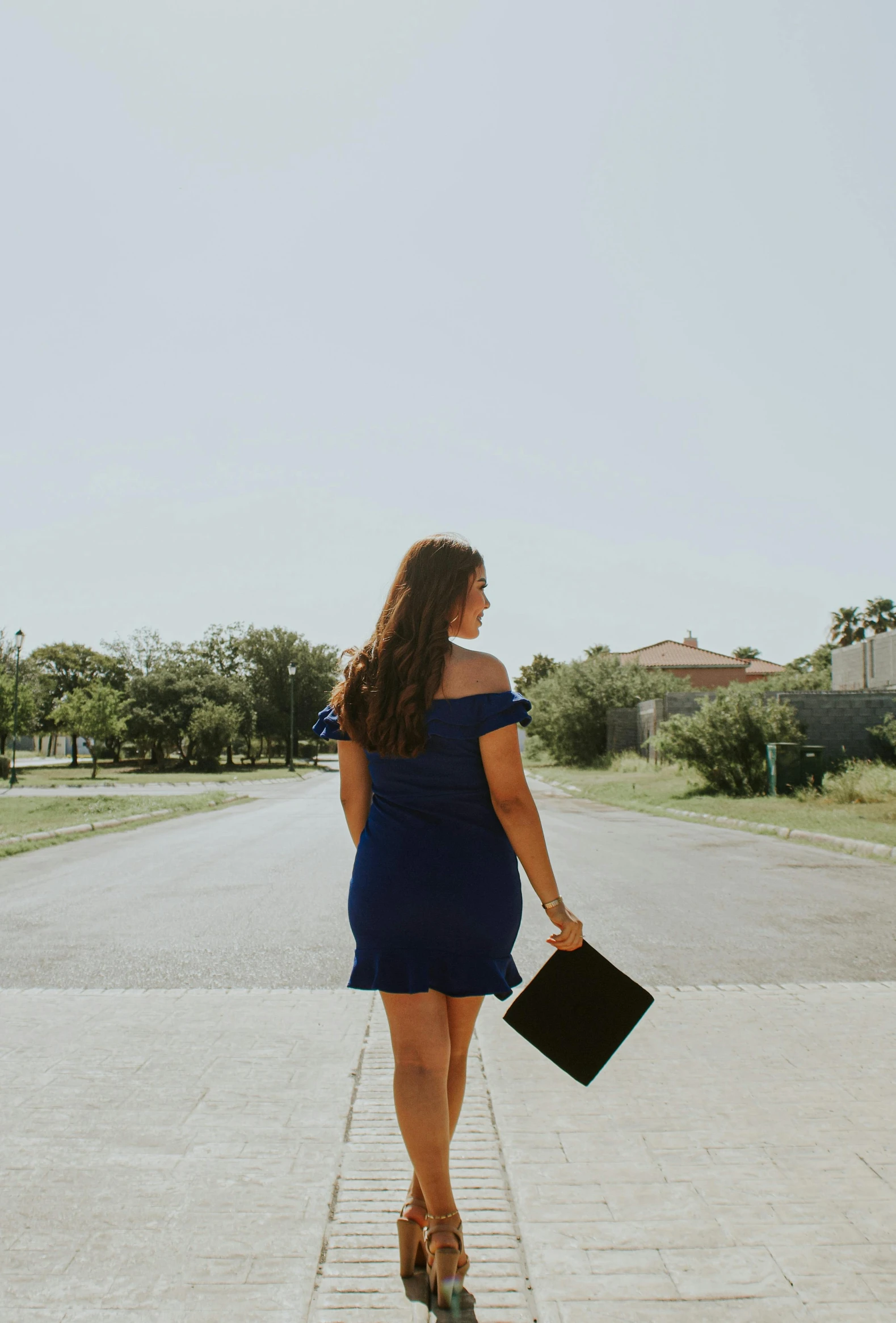 a woman in a blue dress walking down a street, an album cover, unsplash, graduation photo, showing her shoulder from back, in a suburb, carrying a tray