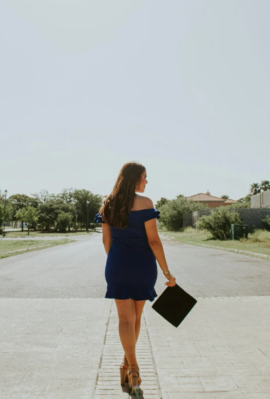 a woman in a blue dress walking down a street, an album cover, unsplash, graduation photo, showing her shoulder from back, in a suburb, carrying a tray
