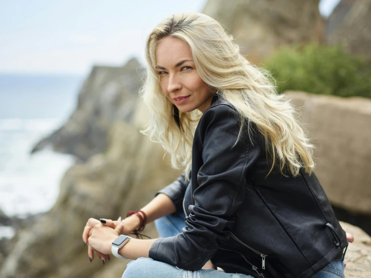 a woman sitting on top of a rock next to the ocean, a portrait, inspired by Károly Lotz, pexels contest winner, wearing a leather flight jacket, long wavy blonde hair, kristanna loken, wearing a jeans jackets