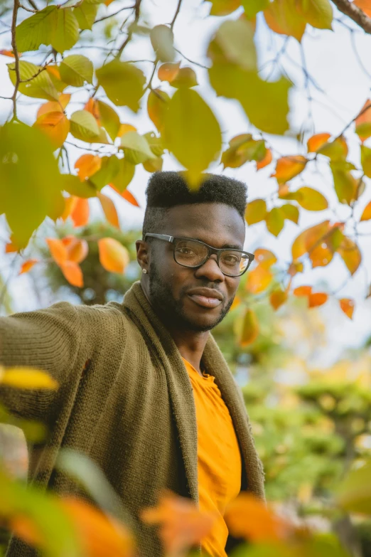 a man with glasses standing under a tree, inspired by Paul Georges, trending on pexels, he is wearing a brown sweater, man is with black skin, fall colors, square rimmed glasses