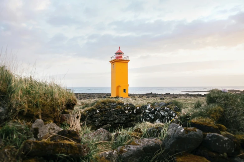 a yellow lighthouse sitting on top of a rocky beach, by Hallsteinn Sigurðsson, gray and orange colours, summer lighting, mustard, brightly-lit