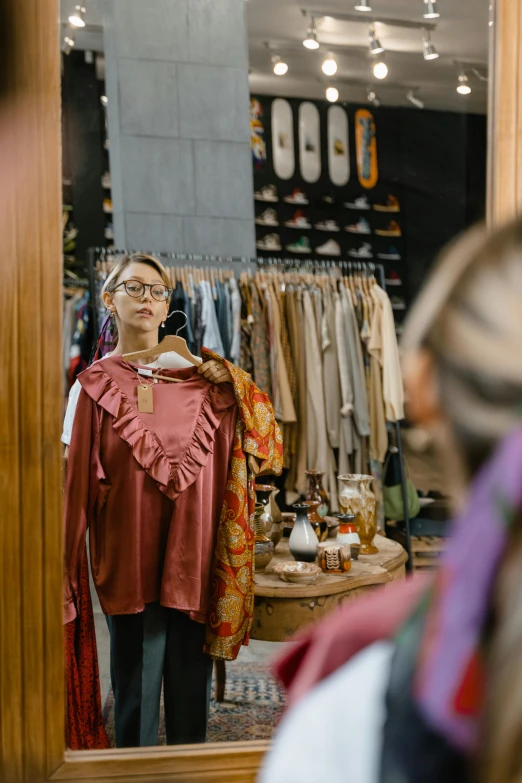 a woman looking at herself in a mirror, trending on pexels, happening, wearing a barca cape, exiting store, brown clothes, inspect in inventory image