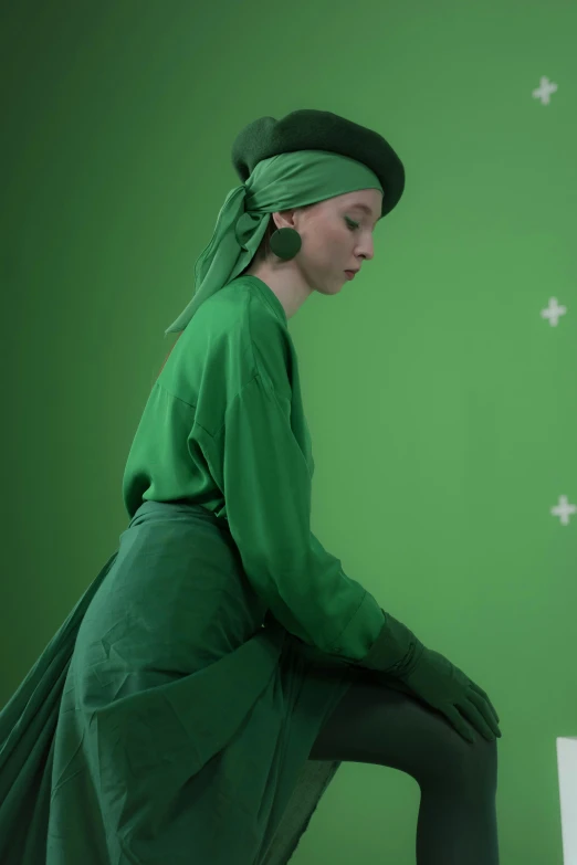 a woman in a green dress sitting on a stool, a colorized photo, inspired by Georges de La Tour, trending on polycount, still from a wes anderson movie, green hat, showstudio, with screens and silks