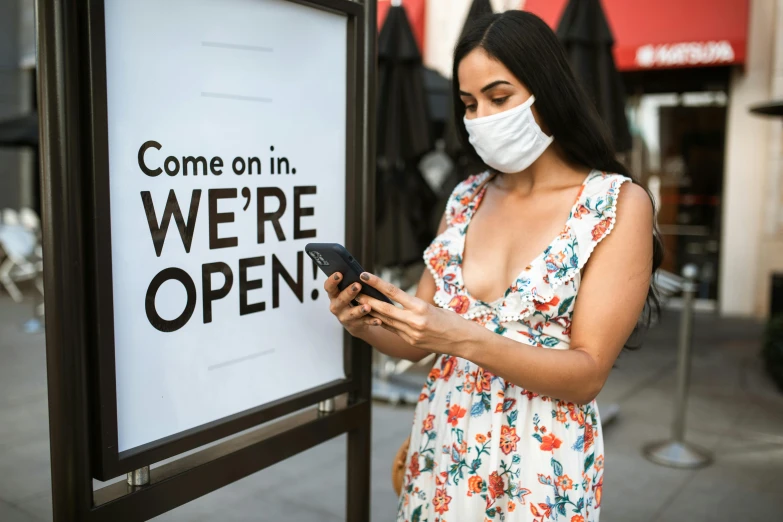 a woman standing in front of a sign that says come on we're open, a photo, trending on pexels, renaissance, surgical mask covering mouth, square, people shopping, checking her phone