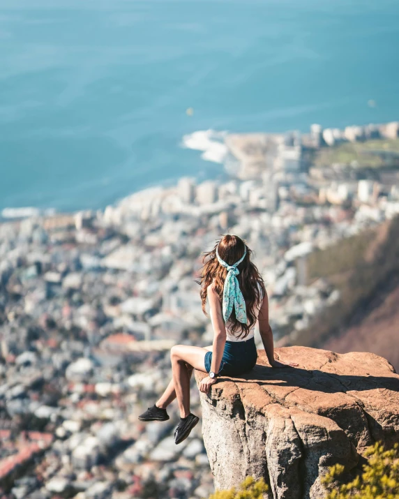 a woman sitting on a rock overlooking a city, pexels contest winner, south african coast, al fresco, girl with plaits, bird's view