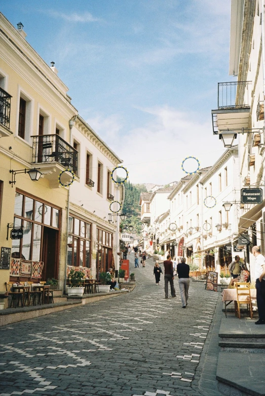 a group of people walking down a cobblestone street, a picture, white buildings, castelvania, restaurant, hillside