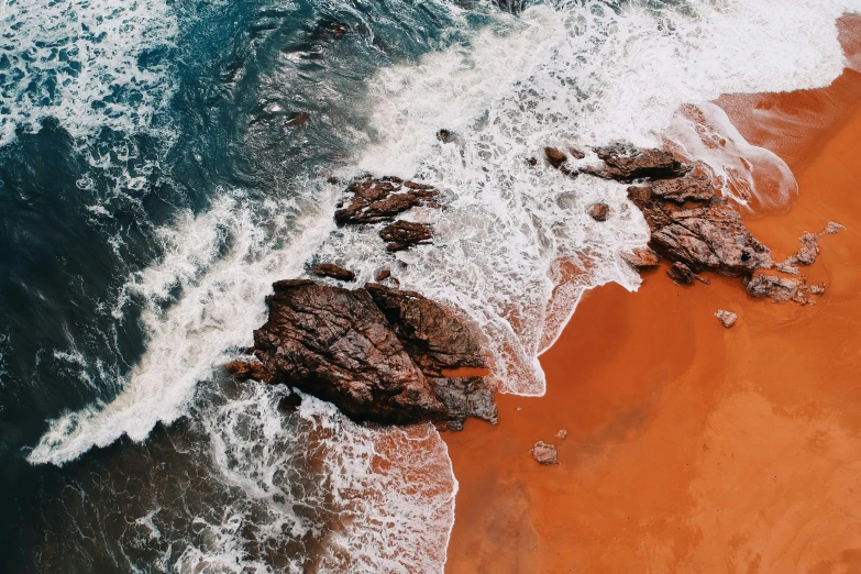 a couple of rocks sitting on top of a sandy beach, pexels contest winner, orange and white color scheme, turbulent water, taken from a plane, red sand