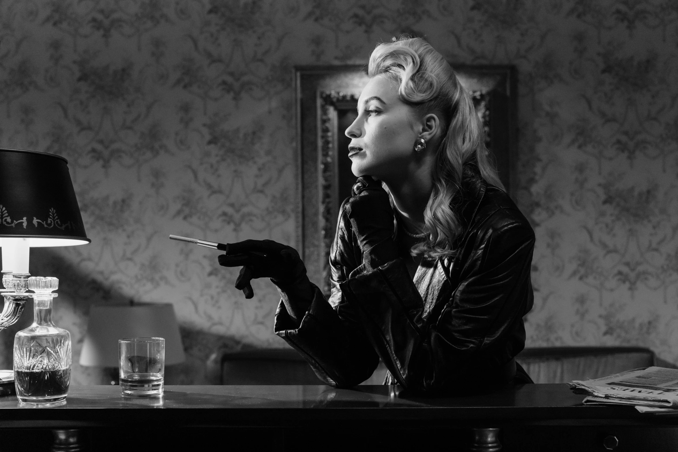a black and white photo of a woman smoking a cigarette, by Emma Andijewska, fantastic realism, in an old 1950s leather jacket, blonde, hand on table, holding a whip