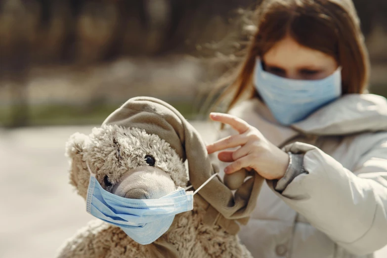 a woman wearing a face mask holding a teddy bear, a picture, by Emma Andijewska, shutterstock, children, game ready, dusty place, urban surroundings