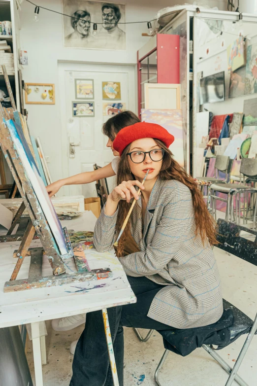 a woman sitting at a table in front of a painting, trending on pexels, academic art, wearing a french beret, holding paintbrushes, girl in studio, tall female emo art student