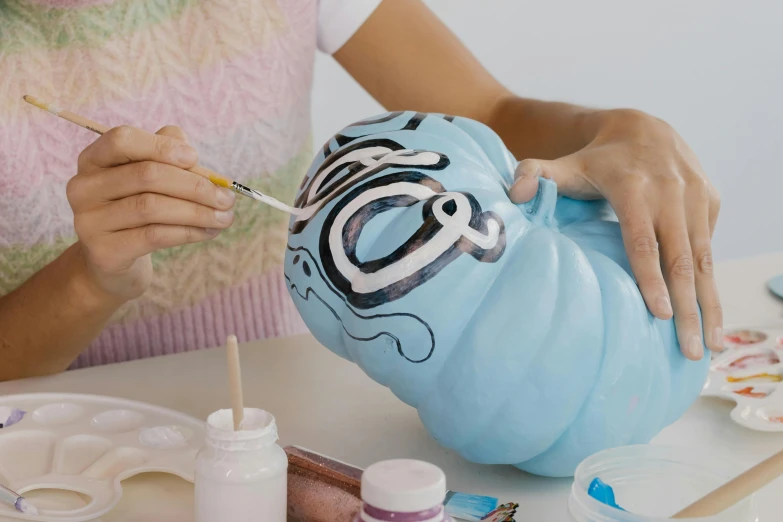 a woman painting a blue pumpkin on a table, inspired by Sarah Lucas, flowing lettering, claymation character, made of cotton candy, on a canva