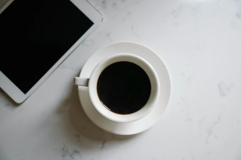 a cup of coffee next to a tablet computer, by Robbie Trevino, pexels contest winner, minimalism, black color on white background, marble table, dark angel of coffee, white