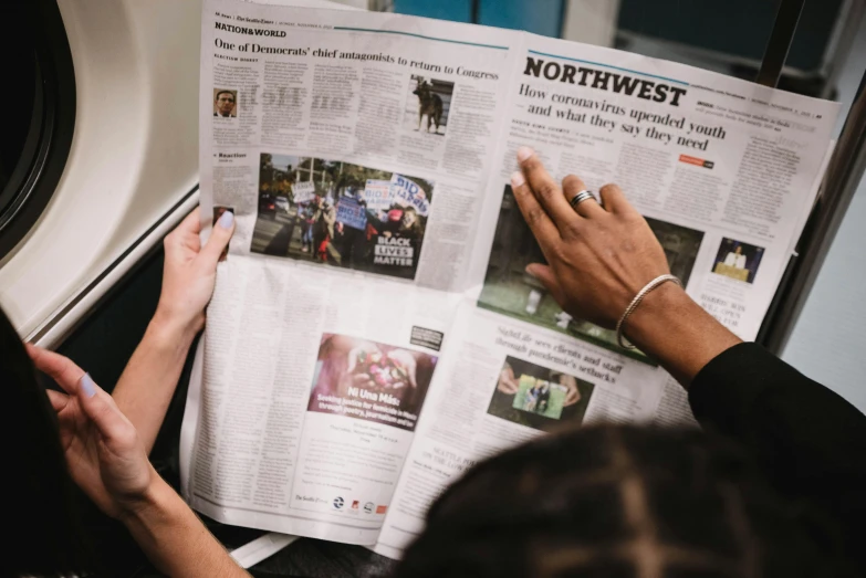 a person reading a newspaper on a train, by Carey Morris, pexels contest winner, holding it out to the camera, diverse, seattle, medium shot of two characters