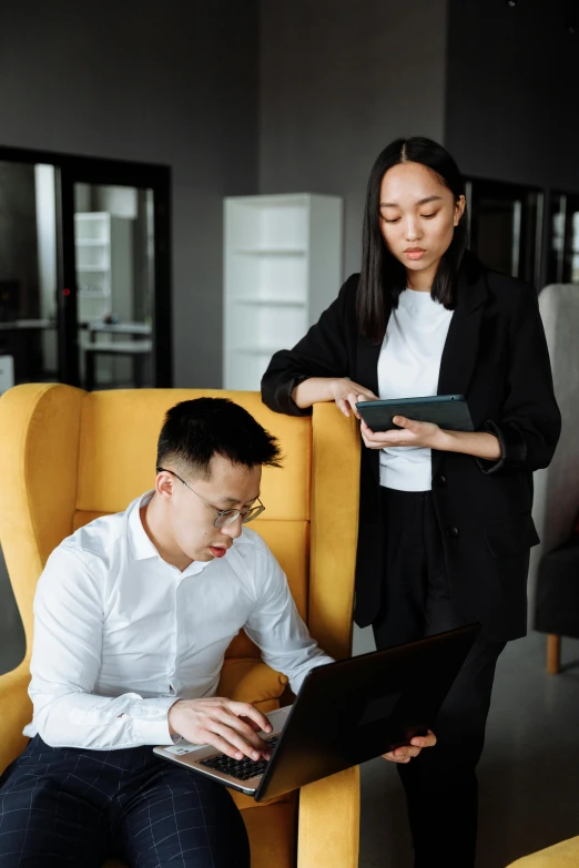 a man sitting in a chair next to a woman on a laptop, asian descent, vp of marketing, yellow and black color scheme, serious business