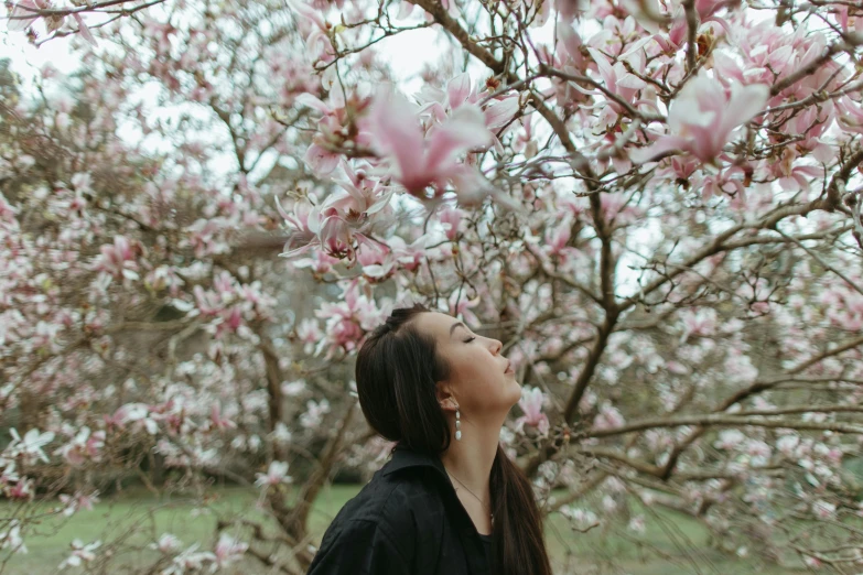 a woman standing in front of a blooming tree, pexels contest winner, magnolias, dreaming of kissing a girl, botanicals, low quality photo