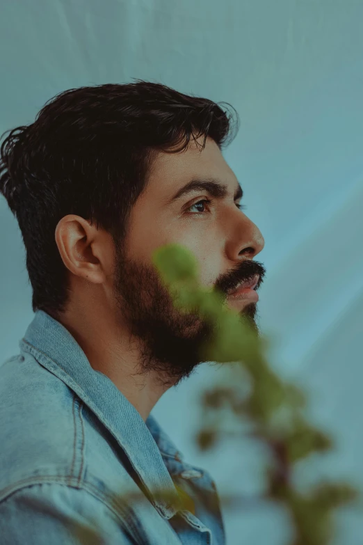 a man with a beard standing in front of a plant, an album cover, by Adam Dario Keel, pexels contest winner, a portrait of rahul kohli, side profile waist up portrait, androgynous person, wearing a light blue shirt
