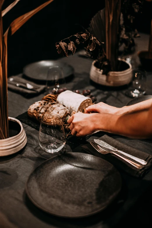 a person setting a table with plates and silverware, charcoal and champagne, holding a baguette, deep texture, food. craft and adventure