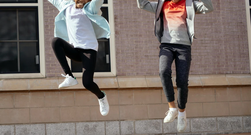two people jumping in the air in front of a building, by Alice Mason, trending on pexels, happening, cute sportswear, background image, school class, product introduction photo