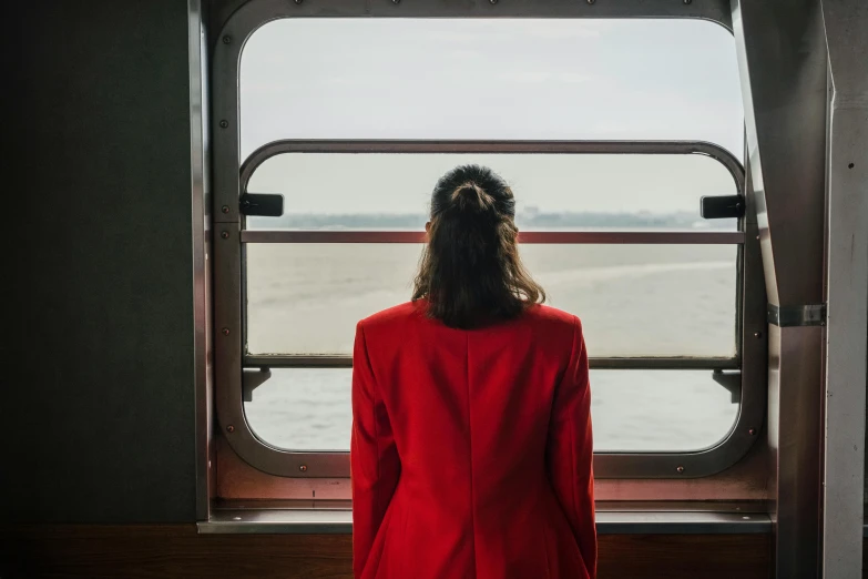 a woman in a red jacket looking out a window, pexels contest winner, on ship, train far, girl in suit, annie lebowitz