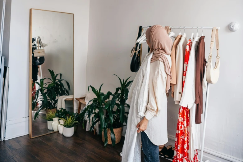 a woman standing in front of a rack of clothes, trending on pexels, hurufiyya, wearing a head scarf, standing in corner of room, foliage clothing, wearing a white robe