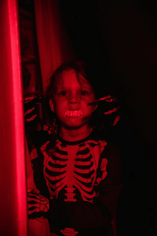 a little girl standing in front of a red light, glowing bones, red facial stripe, showing teeth, against a red curtain