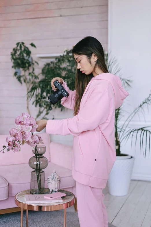 a woman standing in a living room holding a camera, inspired by Ruth Jên, instagram, wearing a pastel pink hoodie, sitting with flowers, in an action pose, product introduction photo