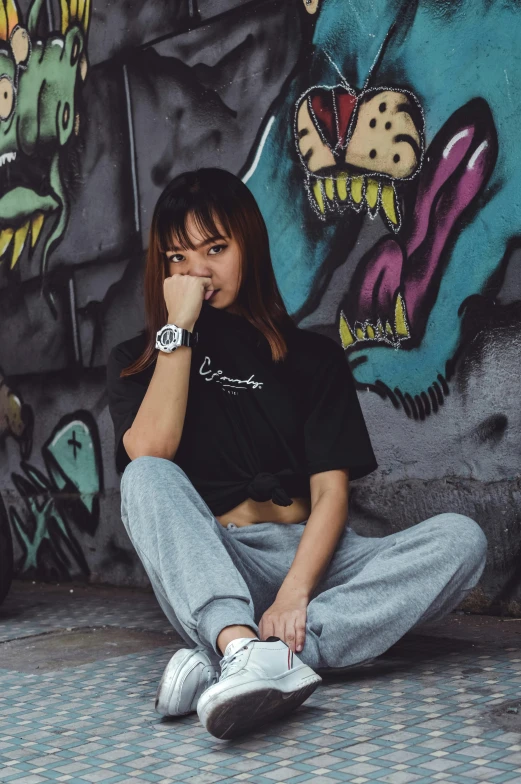 a woman sitting on the ground in front of a graffiti wall, a cartoon, inspired by Wang E, trending on unsplash, with ripped crop t - shirt, wearing a watch, low quality photo, young asian woman